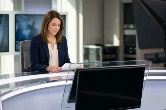 Woman in a blue blazer reading papers at a lit studio desk, with monitors in the foreground and background